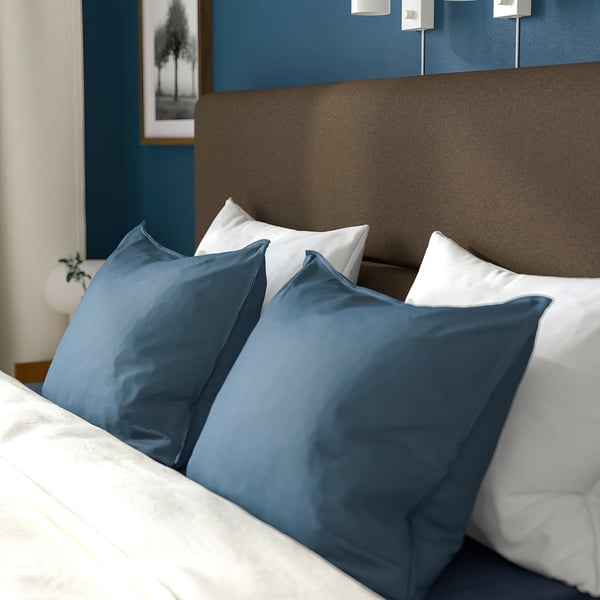 Ein aufgeräumtes Bett mit weichem Kopfteil, weißen und blauen Kissen, das Komfort und Unterstützung beim Lesen oder Fernsehen bietet.