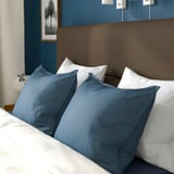 Ein aufgeräumtes Bett mit weichem Kopfteil, weißen und blauen Kissen, das Komfort und Unterstützung beim Lesen oder Fernsehen bietet.