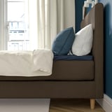 Bett mit weichem Kopfteil für die Rückenlehne, weiße Laken, blaue Kissen, am Fenster.
