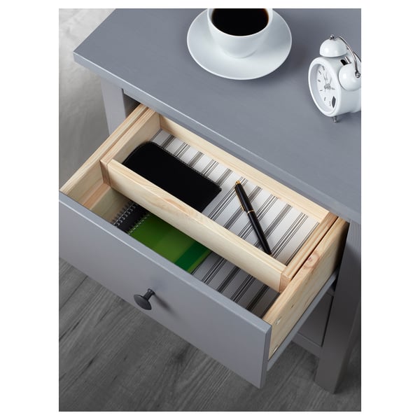 Grauer Nachttisch mit offener Schublade, die Stifte und Notizbuch in einem hölzernen Organizer, Wecker und Kaffee darauf freigibt.