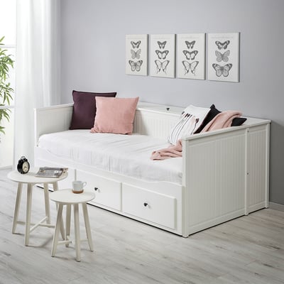 HEMNES weißes Tagesbett mit Stauraum, Kissen und Decke. Kleine Beistelltische mit einer Tasse und einem Buch. Schmetterlingsdrucke an den Wänden.