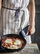 Eine Person mit einer gestreiften Schürze hält eine Pfanne mit Frühstück auf einem rustikalen Tisch.