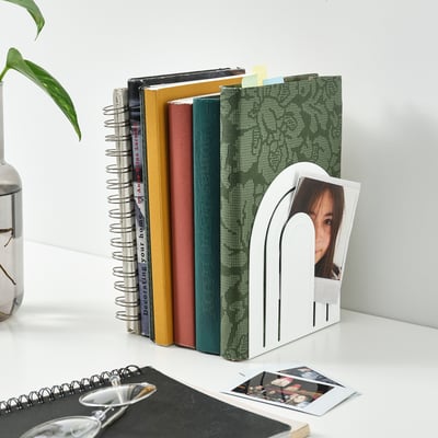 Bücherregal mit weißen HÖSSLA Buchstützen mit bunten Büchern, Pflanzen und Fotos.