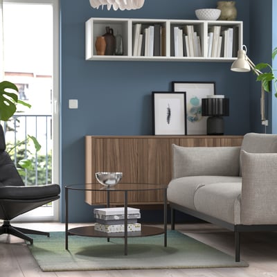 Modernes Wohnzimmer mit dunkelblauen Wänden, hellgrauem Sofa, Couchtisch aus Glas, Holzschrank und einem großen grünen und beigen Teppich mit Farbverlauf.