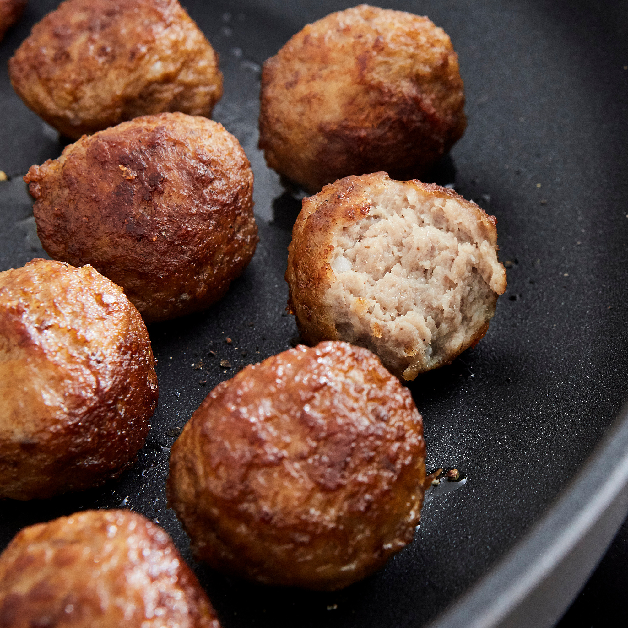 Braune Fleischbällchen, die in einer schwarzen Pfanne gebraten werden, eine in Scheiben geschnitten, um das Innere zu zeigen.