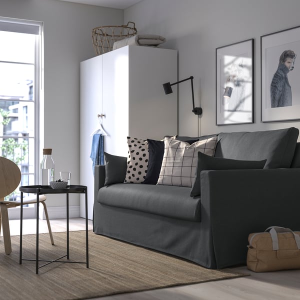 Modernes Wohnzimmer mit grauem HYLTARP-Schlafsofa, karierten/gepunkteten Kissen, Couchtisch mit Flasche/Glas, Hundefotos, weißem Schrank, großem Fenster.