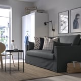 Modernes Wohnzimmer mit grauem HYLTARP-Schlafsofa, karierten/gepunkteten Kissen, Couchtisch mit Flasche/Glas, Hundefotos, weißem Schrank, großem Fenster.