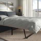 Modernes graues Schlafsofa mit schwarzen Metallbeinen, weichen Kissen, vor grauer Wand mit abstrakter Kunst. Daneben Tisch mit Lampe, großes Fenster.