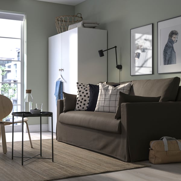Modernes Wohnzimmer mit HYLTARP-Schlafsofa, bunten Kissen, Hundeporträts, Kleiderschrank, Glastisch.