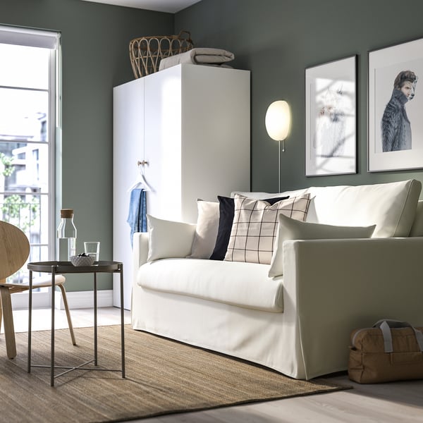 Modernes Wohnzimmer mit HYLTARP-Sofa, Kissen, Beistelltisch und Kleiderschrank vor grüner Wand mit Hundeporträts und Schiebetür.