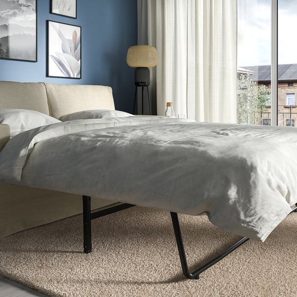 Modernes graues Schlafsofa mit dunklen Beinen in einem gut beleuchteten Raum. Graue Decken und Kissen, weiße Vorhänge, Stehlampe und gerahmte Bilder an der blauen Wand.