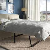 Modernes graues Schlafsofa mit dunklen Beinen in einem gut beleuchteten Raum. Graue Decken und Kissen, weiße Vorhänge, Stehlampe und gerahmte Bilder an der blauen Wand.