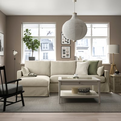 Modernes Wohnzimmer mit cremefarbenem HYLTARP-Sofa, Stauraum und weißem Couchtisch. Große Fenster, beiger Teppich, schwarz/weißer Stuhl und Pendelleuchte.