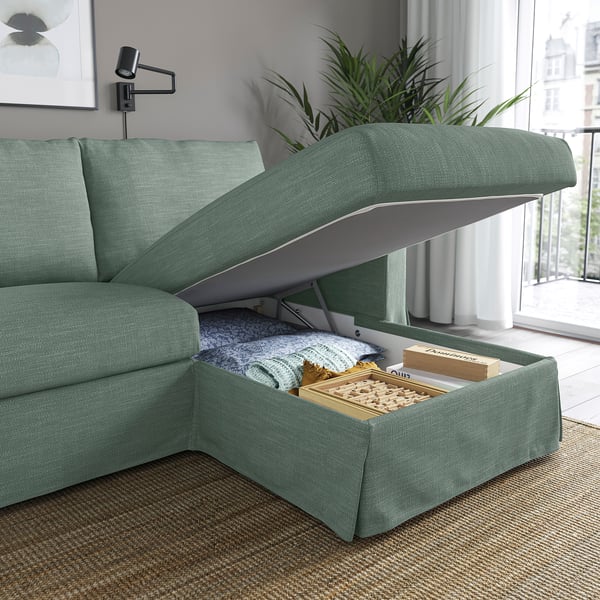 Grünes Sofa mit offenem Staufach unter der Chaiselongue, auf dem eine gefaltete Decke, Bücher und ein Brettspiel zum Vorschein kommen.