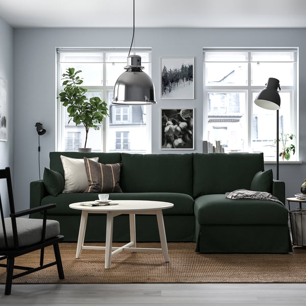 Modernes Wohnzimmer mit großem grünem L-förmigem Sofa, weißem Couchtisch und Pendelleuchte. Große Fenster und Kunstwerke an den Wänden.