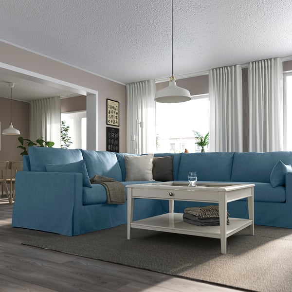 Helles Wohnzimmer mit großem blauem L-förmigem HYLTARP-Sofa, weißem Couchtisch, Hängelampen, Topfpflanzen und großen Fenstern mit Vorhängen.