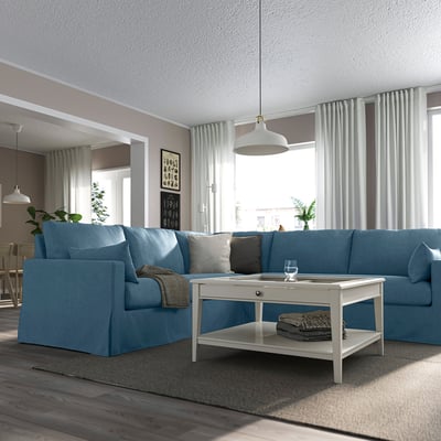 Helles Wohnzimmer mit großem blauem L-förmigem HYLTARP-Sofa, weißem Couchtisch, Hängelampen, Topfpflanzen und großen Fenstern mit Vorhängen.