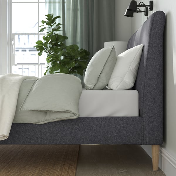 Modernes Schlafzimmer mit grauem Polsterbett, weißen Kissen, grüner Decke, Pflanze und Stehlampe.