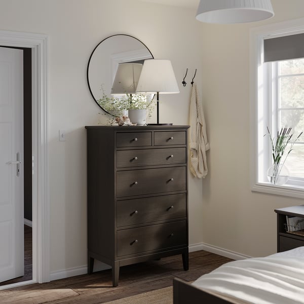 Schlafzimmer mit Kommode aus dunklem Holz, rundem Spiegel, weißer Lampe, hellgelben Wänden.