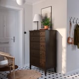 Moderner Eingangsbereich mit brauner IDANÄS-Truhe, weißer Lampe, grüner Pflanze, Kleiderhaken mit Jacken, schwarz-weißem Fliesenboden und Korbstuhl.