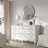 Schlafzimmer mit weißer IDANÄS-Kommode, rundem Spiegel, Lampe und Vase mit Blumen.
