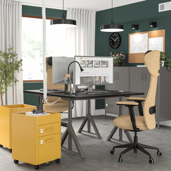Modernes Büro: schwarzer IDÅSEN-Schreibtisch, ergonomischer beiger Stuhl, gelber Aktenschrank. Grüne Wände, weiße Vorhänge, Uhr. Geräumig, gut organisiert.