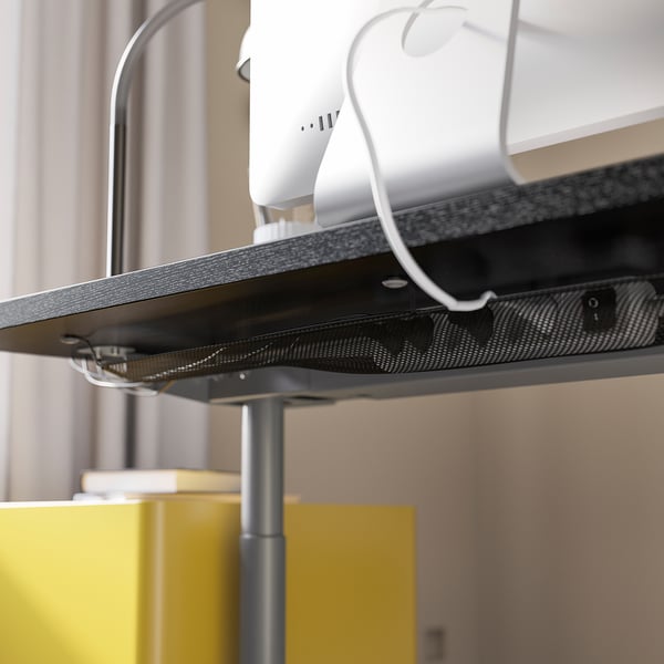 Ein schwarzer IDÅSEN-Schreibtisch mit einem Netzkabelführungsnetz darunter, das ein weißes Gerät mit einem sichtbaren Kabel trägt.