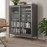 Schwarzer IDÅSEN-Schrank mit Glastüren und Metallrahmen, in dem Bücher und Dekoration Platz finden, höher als Stuhl und Pflanze in der Nähe.