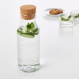 Karaffe aus klarem Glas mit Korkverschluss, die Wasser und Minzblätter enthält. Positioniert in der Nähe von zwei Gläsern und Keksen auf weißer Fläche.