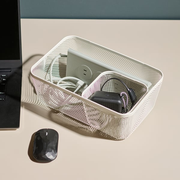 In einem Mesh-Aufbewahrungsbehälter auf einem Schreibtisch befindet sich technisches Zubehör, darunter Kabel und ein Ladegerät, neben einem Laptop und einer Maus.