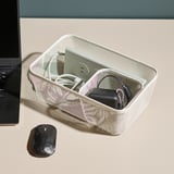 In einem Mesh-Aufbewahrungsbehälter auf einem Schreibtisch befindet sich technisches Zubehör, darunter Kabel und ein Ladegerät, neben einem Laptop und einer Maus.