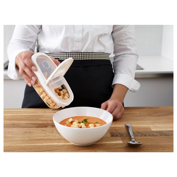 Person, die Croutons aus einem transparenten IKEA 365+ Glas in eine Schüssel mit Suppe auf einer Holzoberfläche gießt. Ein Löffel ist in der Nähe.