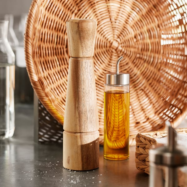 Pfeffermühle aus Holz mit Keramikmahlwerk auf dem Tisch, neben einer geflochtenen Platte und einem Ölspender.