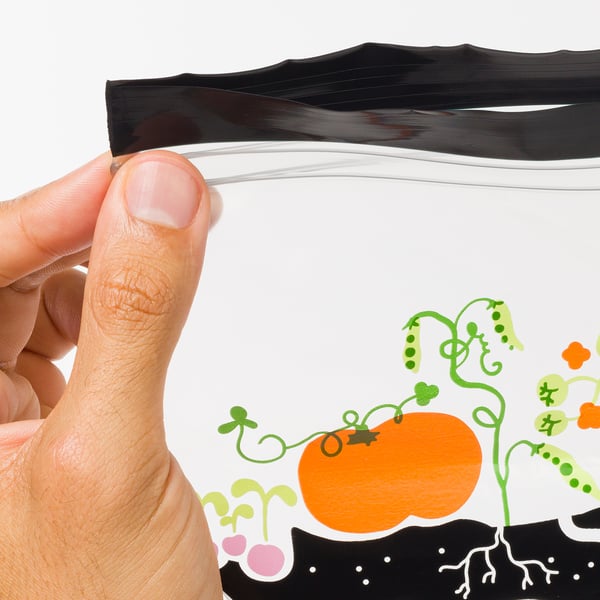 In der Hand hält er eine offene, wiederverwendbare Aufbewahrungstasche mit bunten Gemüseillustrationen, die die dauerhafte Dichtigkeit und Qualität betonen.