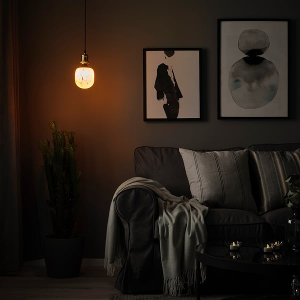 Gemütliches Wohnzimmer mit dunklen Möbeln, warmer Lampe, grauer Couch, Wandkunst, Pflanze und Kerzen.