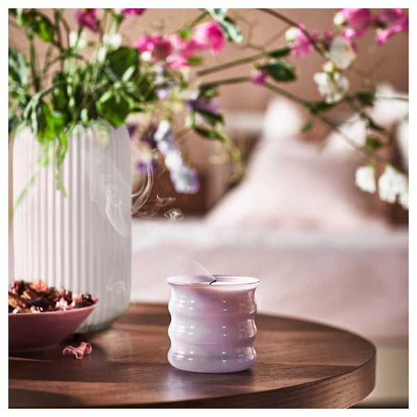 Lila-graue Duftkerze, JÄMNMOD, auf Holztisch mit rosa Blumen in der Vase.