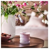 Lila-graue Duftkerze, JÄMNMOD, auf Holztisch mit rosa Blumen in der Vase.