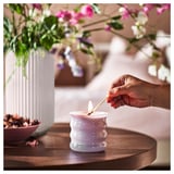Stellen Sie sich eine Hand vor, die eine weiße JÄMNMOD-Duftkerze in einem strukturierten Glasbehälter auf einem Tisch neben einer Vase mit bunten Blumen anzündet.