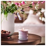 Eine brennende weiße JÄMNMOD-Duftkerze auf einem Holztisch, neben einer Vase mit rosa und lila Blumen.