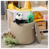 Großer grauer JÄRRESTAD Hocker zur Aufbewahrung von Spielzeug im Zimmer mit Stofftieren: Panda, Dinosaurier, Außerirdischer und Partyzubehör.