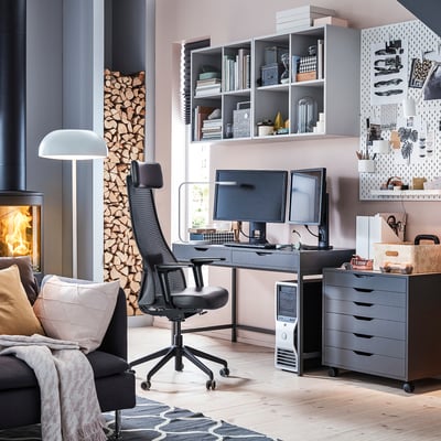 Home-Office-Einrichtung mit ergonomischem schwarzem Stuhl, Schreibtisch, zwei Monitoren, Kamin, Regalen und Dekor.