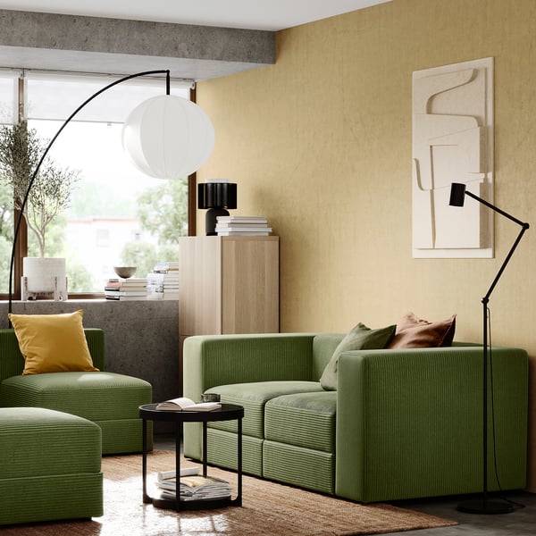 Modernes Wohnzimmer mit grüner JÄTTEBO-Sofagarnitur, gelben Kissen, schwarzer Stehleuchte und beiger Wandkunst.