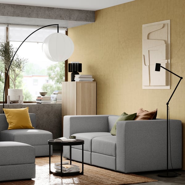 Modernes Wohnzimmer mit grauem modularem JÄTTEBO-Sofa, gelben Kissen, Stehlampe, Beistelltisch, Bücherregal mit Büchern und abstrakter Wandkunst.