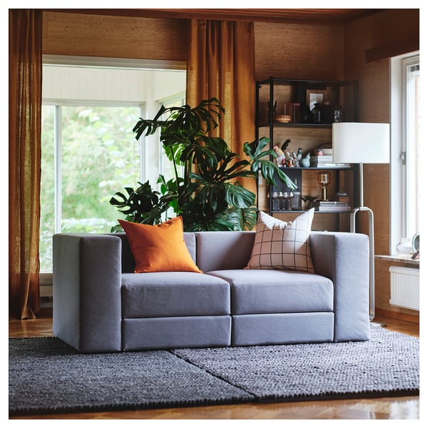 Modernes Wohnzimmer mit grauem modularem JÄTTEBO-Sofa, orangefarbenen Kissen, großer Pflanze, Holzböden und Vorhängen.