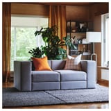 Modernes Wohnzimmer mit grauem modularem JÄTTEBO-Sofa, orangefarbenen Kissen, großer Pflanze, Holzböden und Vorhängen.