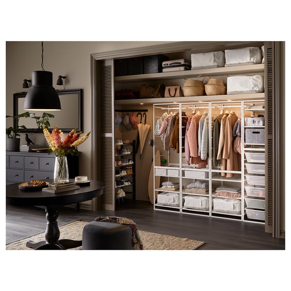 Gut organisierter Kleiderschrank mit weißem JONAXEL-Aufbewahrungssystem, hängender Kleidung, Regalen mit Körben und einem Beistelltisch mit Blumen und Büchern.