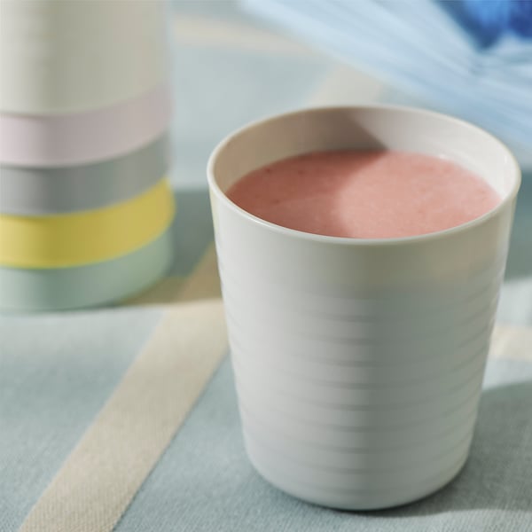 Eine weiße Babytasse sitzt auf einem gestreiften Tuch, in dem sich ein rosa Smoothie befindet. Dahinter sind bunte stapelbare Becher zu sehen.