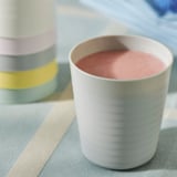 Eine weiße Babytasse sitzt auf einem gestreiften Tuch, in dem sich ein rosa Smoothie befindet. Dahinter sind bunte stapelbare Becher zu sehen.