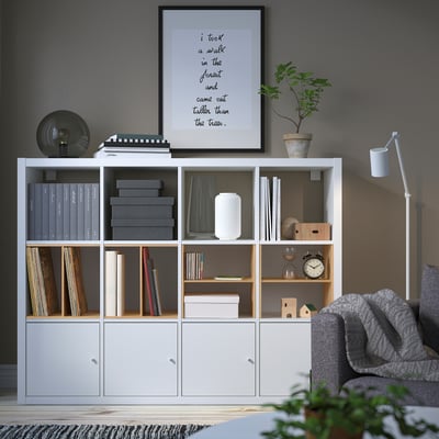 Modernes Wohnzimmer mit KALLAX Regal gefüllt mit Büchern, Dekor und Pflanzen. Graues Sofa mit Decke, gemütlich und organisiert.