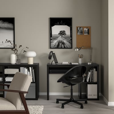 Moderne Home-Office-Einrichtung: schwarzer Schreibtisch mit Laptop, Lampe und Stuhl. Schwarz gerahmte Kunst auf grauen Wänden. Beige Stuhl und Lampe im Bücherregal.